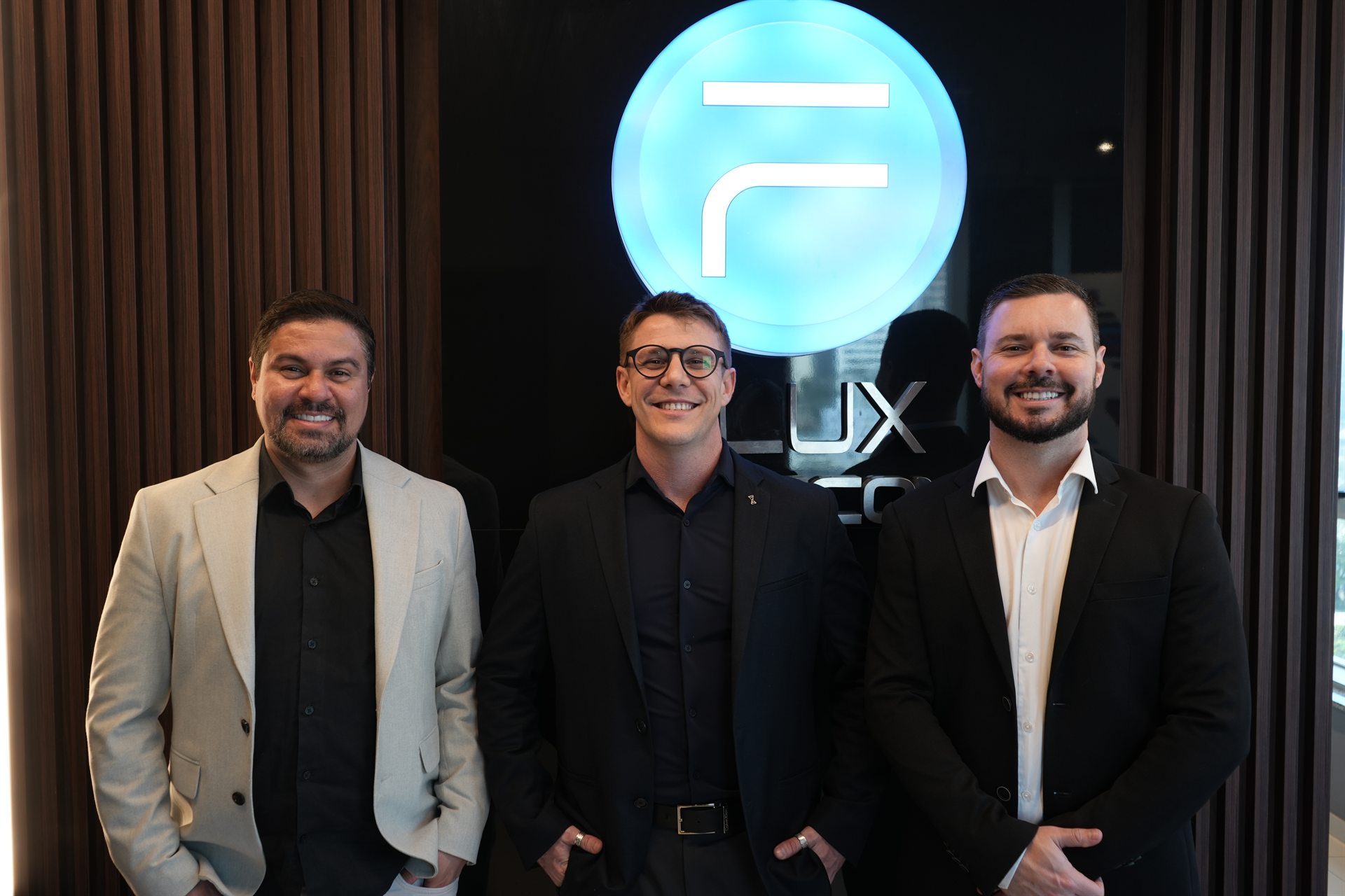 Os três diretores da Flux Tecnologia lado a lado na recepção da empresa, sob o símbolo iluminado da marca.